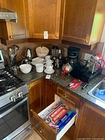 Corner kitchen countertop showing coffee maker, two French presses, coffee cups and dishes, decanters, bowls, and storage drawer with parchment and cling wrap.