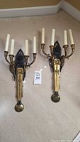 Pair of brass wall sconces laid flat, front view showing four arms with faux candle sleeves, decorative central figure and bottom finials