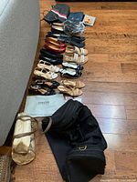 Wide view of assorted women's designer shoes in various styles and colors on wooden floor next to a gray couch, alongside several handbags including a metallic gold bag and a black crossbody purse.