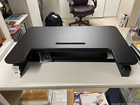 Full view of black adjustable desk riser on white desk with brown office chair and office supplies in background.