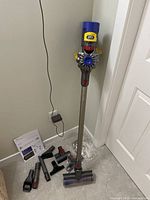 Assembled vacuum standing upright next to door with charger plugged in and attachments laid out on floor.