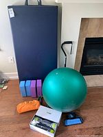 Photo showing dark blue floor mats, green yoga ball, resistance bands, Zumba shake weights, and walking cane on wooden floor near fireplace.