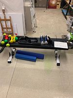 Wide view showing black flat exercise bench with silver legs, an assortment of dumbbells, four grip balls, two shake weights, and workout program chart on the bench.