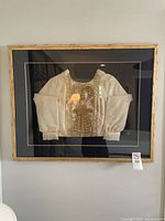 Framed white shirt with gold embroidery and pearl beadwork, centered in black matte and in a gold-painted wood frame showing wear.