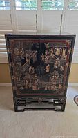 Full view of black lacquered wood cabinet with hand-painted Chinese palace and figure scenes, brass hardware, and stand.