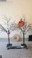 Two artificial LED blossom trees standing on black square bases, with a decorative white wreath and orange extension cord behind clustered against a plain wall.