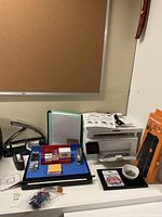 Desk surface with HP LaserJet Pro MFP printer, Fiskars SureCut trimmer box, adjustable laptop stand, stapler, hole punch, stationery organizer, push pins and binder clips in bags, and small silver bowl with miscellaneous items.