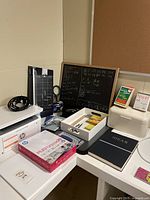 Photo showing the entire lot arranged on white desks including printer, paper ream, chalkboard, corkboard, sharpener, paper cutter, storage box, notebook, and chalk.