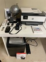 Photo of HP OfficeJet 8015e printer, photo paper, multiple audio recording devices, globe, Apple TV remote, iPad case, power strip, and miscellaneous electronics on a white desk and underneath it.