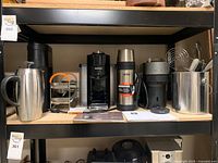 Shelf with two Nespresso Vertuo coffee makers, a large thermos, Kcup pod holding rack, and Kcup throw bin with kitchen utensils.