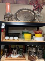 Shelf view showing large hurricane candle vase on top with large silver platter behind it; below shelves holding yellow ceramic bowls, white bowls, green lidded container, and small house spice rack; bottom shelf with coffee press, cookie jar, cheese grater, and serving trays.