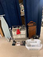 Photo showing rolling laundry hamper with three fabric compartments, ironing board with cover, two irons, suit butler with wooden panel and trouser press, and three white plastic laundry baskets.