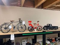 Photo showing three miniature models displayed on a shelf including a black vintage bicycle, red rickshaw tricycle, and black 1940 Ford pickup model, all with store tags attached.