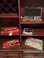 Photo showing wooden shelf with a wooden poker chip set open on middle shelf, containing poker chips and two decks of cards. Next to it, a black and white chess set in a flat box. Below on the lower shelf two red Scrabble game boxes, one unopened. Also on the bottom shelf, a near-new Monopoly Classic Edition board game in green and white box.