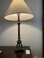 Full view of the lit vintage brass table lamp sitting on a dark wooden surface, showing the beige fabric shade and patina on the metal base.