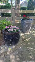 Two planters placed outdoors on paving stones near a wooden fence; the tall gray planter with red flowers is behind the round dark glazed planter with pink flowers and rim chips.