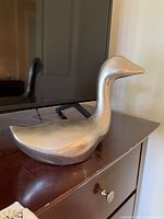 Side view of smooth, solid metal bird sculpture showing elongated neck and rounded body on dark wooden furniture.