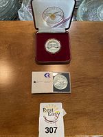 Two silver coins displayed on a wooden surface, one in a maroon presentation box and the other in a protective sleeve with Royal Canadian Mint cardboard insert. A lot number card with '307' is also visible.