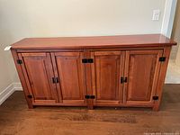 Front view of a solid wood sideboard cabinet with four paneled doors and black metal hinges and handles, placed on a wooden floor against a beige wall.