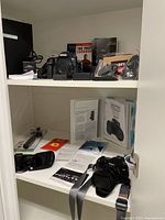 Cabinet interior showing upper shelf with Nikon D850 and books, lower shelf with Sony DSC-RX10M, manuals and accessories.