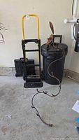 View of yellow and black standup dolly, black collapsible cart, large black garbage bin, and decorative welded snake hook in garage setting.