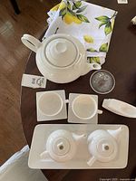 Full assortment on table showing large teapot, two smaller teapots, two cups with square saucers, glass dish with lid, spoon rests, and lemon print tablecloth.