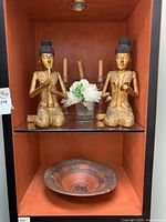 Pair of gilded Thai musician statues kneeling on shelf with clay bowl below and floral decor behind.