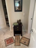 Wooden side table with two drawers, brass and enamel dog sculpture on top, green collectible plate, and embroidered fabrics on the floor.