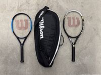 Two Wilson tennis rackets and a black Wilson carry bag arranged on a gray concrete floor