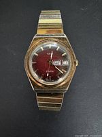 Front view of men's vintage Timex Electro watch showing red dial, gold-tone markers, hands, and day-date window with metal expandable band.