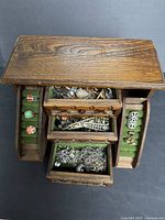 Overhead photo of the wooden jewelry box open, showing multiple drawers and green velvet-lined compartments filled with costume jewelry including rings and brooches.