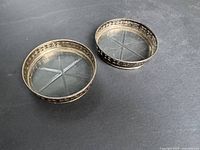 Full view of two round vintage sterling silver coasters with rims featuring floral and geometric cutout designs, glass inset bases showing star pattern.