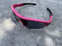 Angle view of pink framed Radar sunglasses with dark wrap-around lens and black temple tips on concrete background.