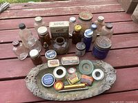 Full lot of antique and vintage medicine and apothecary bottles and tins arranged on an ornate silver metal tray and surrounding area, showing various bottle shapes, sizes, and colors including clear, amber, and cobalt blue glass bottles along with an assortment of metal tins.