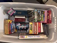 Top-down view of a plastic bin containing multiple vintage board games including Yahtzee, Scrabble, Tic Tac Total, and card decks.