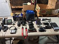 Wide view of all items laid out on table including cameras, phones, cassette players, speakers