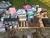 Wide view of large quantity of mixed media on a pallet outdoors including boxes of VHS, cassettes, 8-tracks, records, and a vintage clock and suitcase.