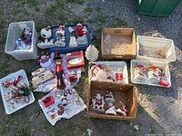 Wide view of assortment of Christmas figurines, plates, and decorative items in boxes and bins arranged on ground outdoors