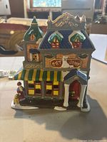 Front view of ceramic light-up Christmas village pet store with figurine and green/yellow awning, showing detailed paint and snow-covered roofs.