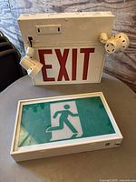 Photo of two exit signs: top sign with red EXIT letters and two spotlights (one broken), bottom sign is green emergency exit running figure sign in white frame.