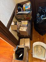 Multiple boxes filled with VHS tapes, fitness equipment, and electronic health devices visible on hardwood floor near wall corner