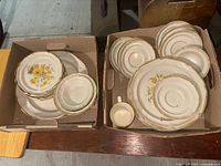 Two boxes containing various plates, cups, and saucers with floral design and gold trim.