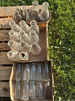 Top view of three cardboard boxes containing different shapes and sizes of glass milk bottles outdoors on grass.