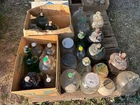 Photo showing multiple large glass jugs of varying colors and sizes arranged on wooden pallets and inside cardboard boxes outdoors in sunlight.