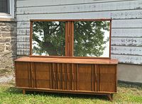 Long wooden dresser with multiple cabinet doors, vertical wood grain patterns, and tapered legs. Large wooden-framed mirror sits behind the dresser, both visible outdoors in natural light.