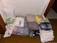 Shows the full assortment of linens including pillow cushions, towels, tablecloths, quilts, and sheets arranged on a carpeted floor against white paneled wall.