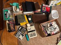 Overview of various office supplies including multiple binders, calculators, small white garbage can, and boxed items containing stationery and other office materials on wooden floor.