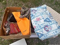 Photo of a cardboard box with assorted fabric pieces and a folded patterned fleece blanket on a white surface outdoors