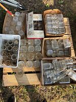 Overview of multiple square glass mason jars in various sizes arranged in boxes and containers on pallet outdoors, some dirt on jars.