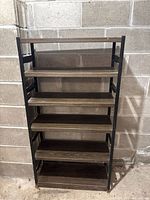 Front view of tall brown metal shelf against a concrete block wall, showing five wooden shelves within a metal frame.